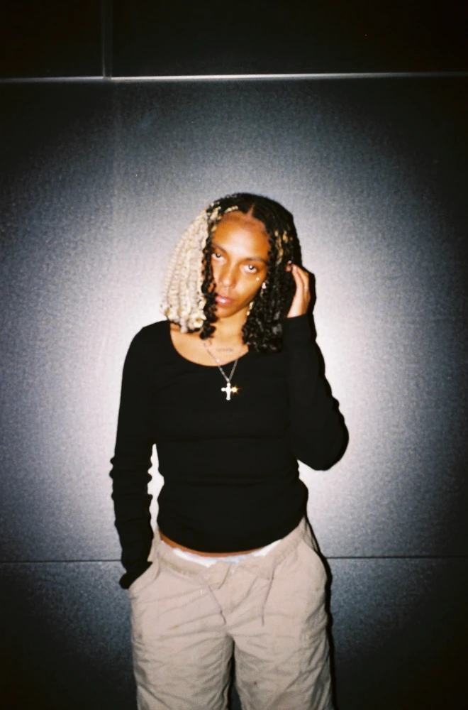 Person with braided hair and a cross necklace, wearing a black top and light-colored pants, standing against a dark background with a vignette effect.