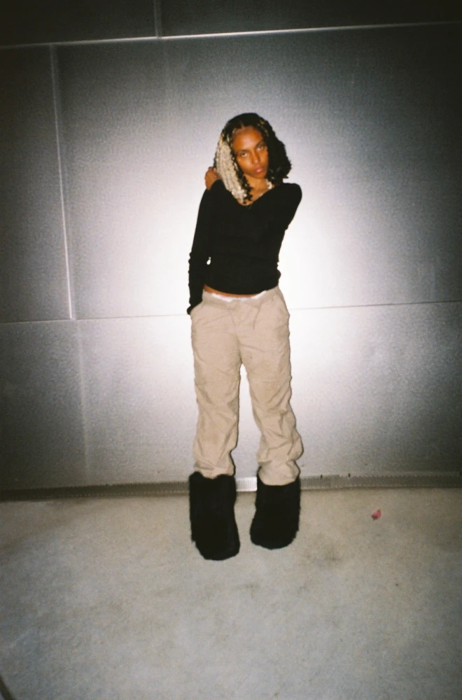 A person standing against a light wall, wearing a black top, beige wide-leg pants, and large fuzzy black boots, posed with a hand near their head and a slightly tilted stance.