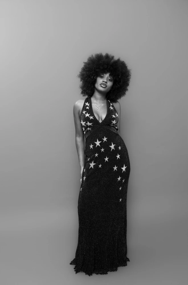 Black and white portrait of a woman with voluminous curly hair wearing a long, flowing black dress adorned with white star patterns, standing confidently against a neutral background.