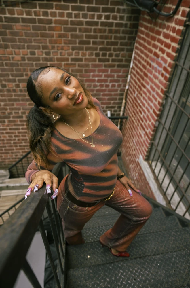 Young woman in striped brown top and matching pants leaning on metal staircase railing against a brick wall background, posing with a confident, relaxed stance.