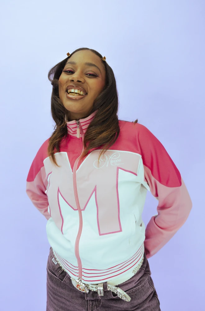 Young woman in a pink and white tracksuit jacket, laughing with joy against a soft purple background, wearing gray jeans and radiating infectious happiness.