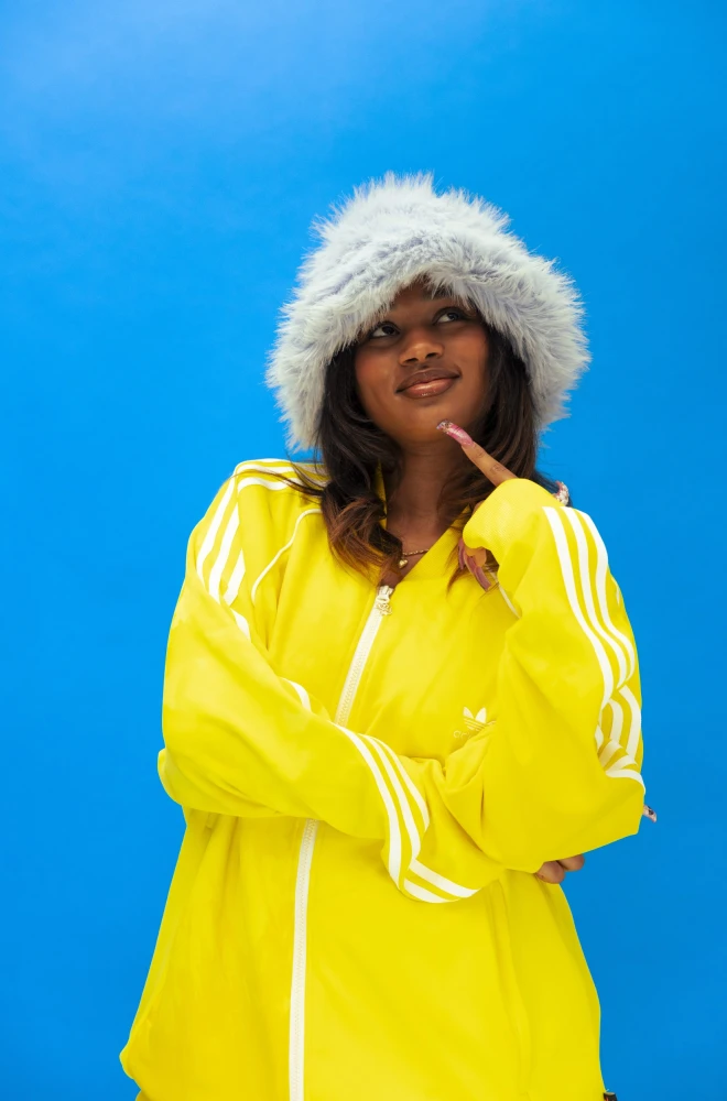 Smiling person in a bright yellow tracksuit and fluffy white winter hat, standing against a vivid blue background with a confident, relaxed pose.