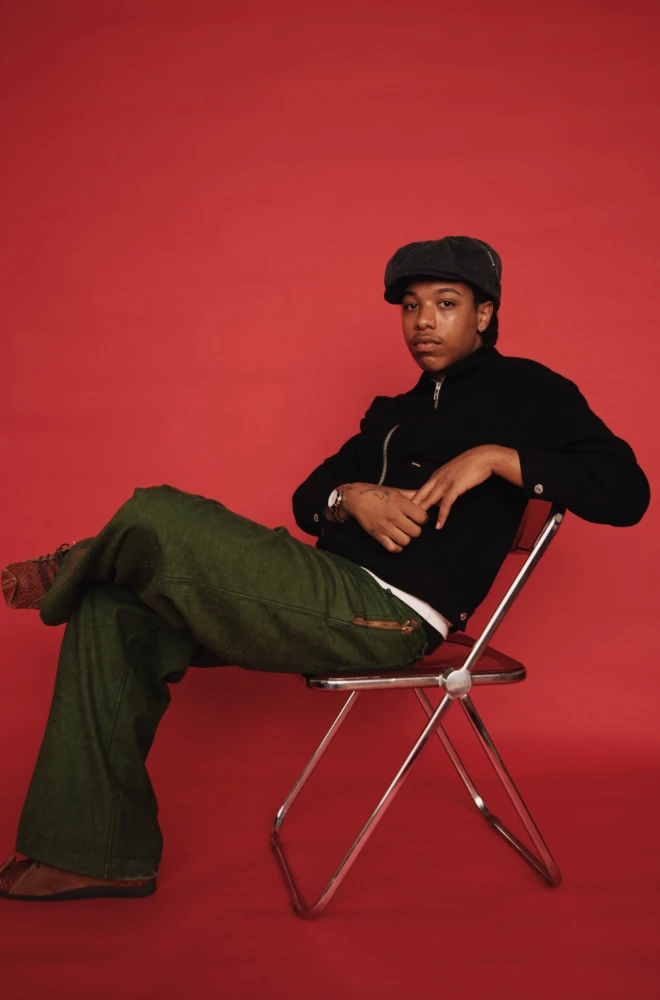 Young person in a black sweater and cap sits casually on a silver folding chair against a red background, wearing green wide-leg pants and leaning back with a relaxed pose.