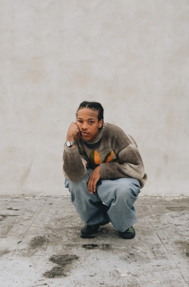 A young person crouches on a concrete surface, wearing a textured brown sweater and blue-gray pants. They rest their head on their hand, looking contemplative against a light, neutral background.