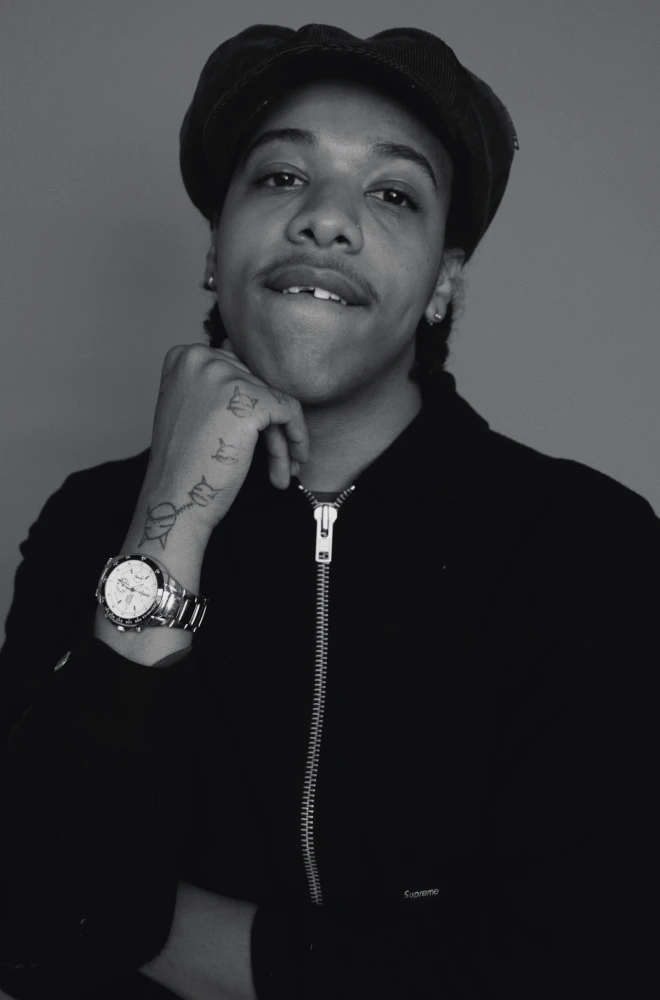 Young person in black jacket and cap, resting chin on hand, wearing a wristwatch, smiling confidently in a black and white portrait photograph.