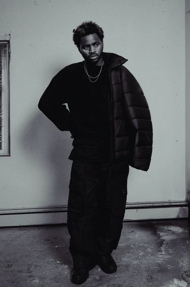 Person in black oversized clothing and puffer jacket standing confidently against a plain wall, with a window and baseboard heater in the background. Moody black and white portrait with dramatic lighting and styling.