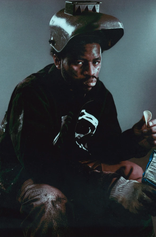 Dark-clad figure wearing an unconventional metallic hat, holding a blue bottle and sitting in a moody, shadowy environment with an intense, contemplative expression.