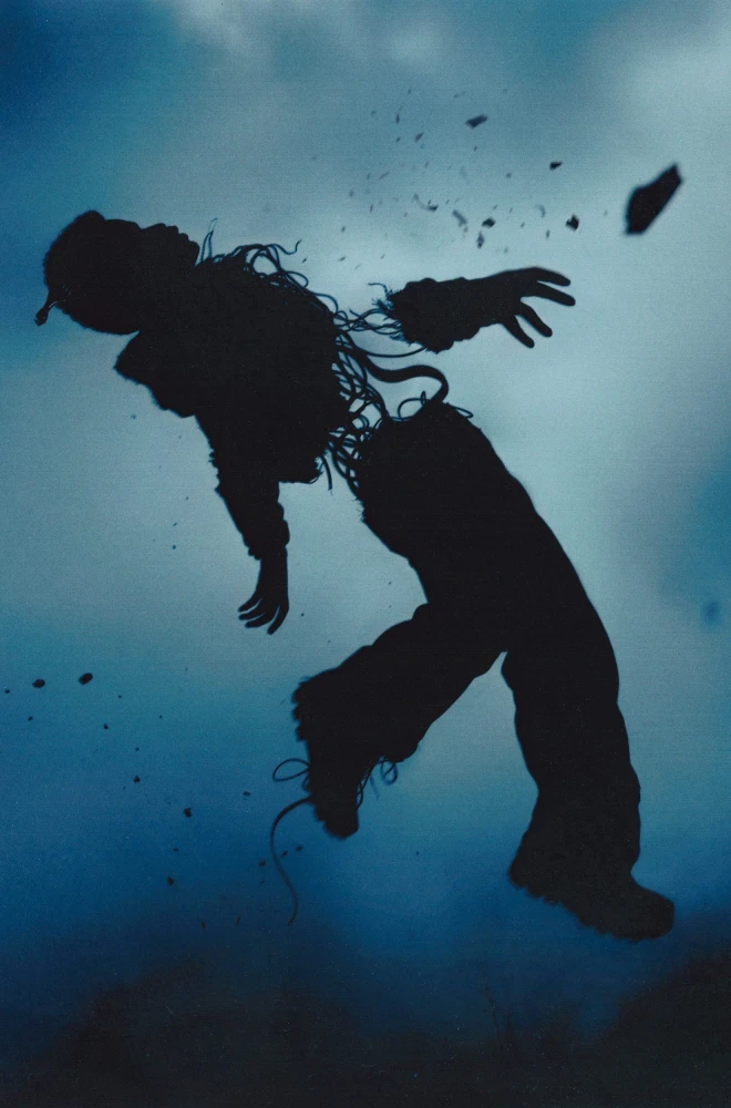 Silhouetted figure leaning back dramatically against a deep blue cloudy sky, with fragments and debris scattering around their dynamic pose, suggesting intense movement or impact.