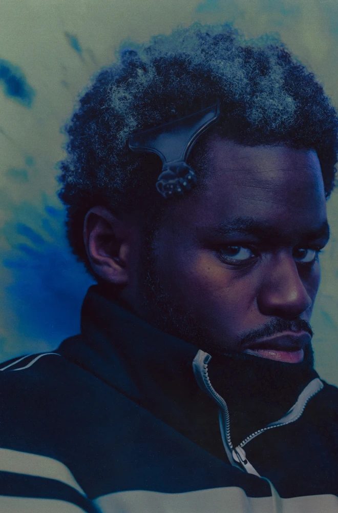 Profile of a young man with curly hair, looking pensively to the side, wearing a black and white striped track jacket against a blue-green textured background.