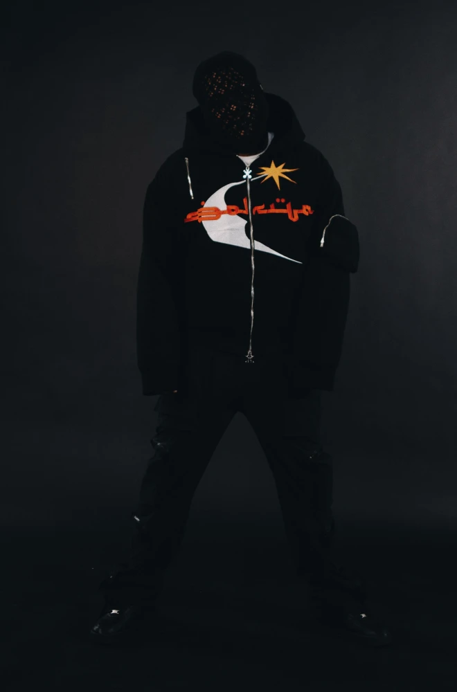 A shadowy figure stands in darkness wearing a black hoodie with a graphic design featuring red and yellow elements. The silhouette is centered against a dark background, creating a moody, mysterious atmosphere.