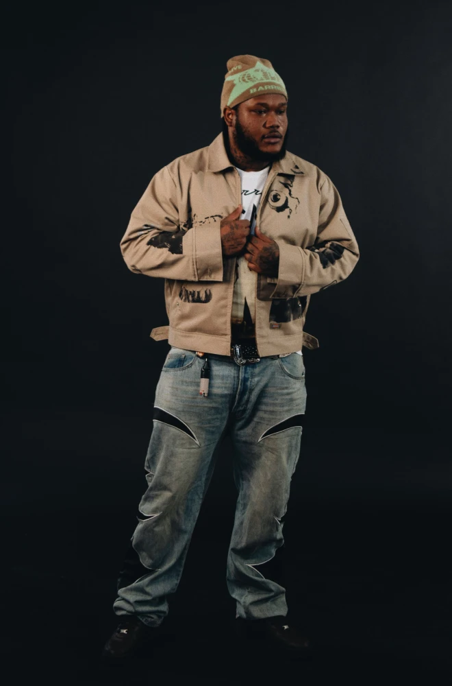 Person wearing a beige workwear jacket, light-wash baggy jeans, and a knit beanie, standing against a black background in a confident pose.