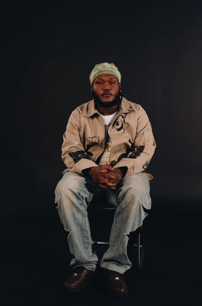 A person sits centered on a chair against a dark background, wearing a beige jacket, light blue jeans, and a knit cap, with hands clasped together.