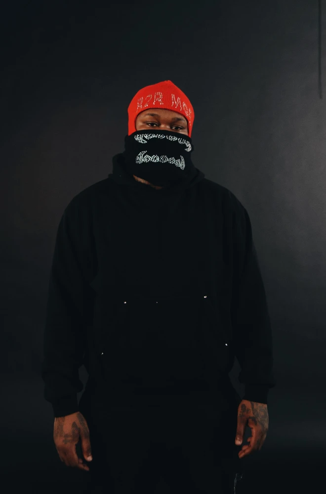 Person wearing a red cap and black bandana covering their face, standing in a dark, shadowy environment with a black coat, creating a mysterious and intense atmosphere.