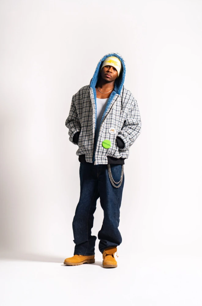 Young person standing against a white background, wearing a gray plaid hooded jacket, blue jeans, and yellow work boots. They have a blue hoodie visible underneath the jacket and a green item in their hand, with a chain hanging from their jeans.