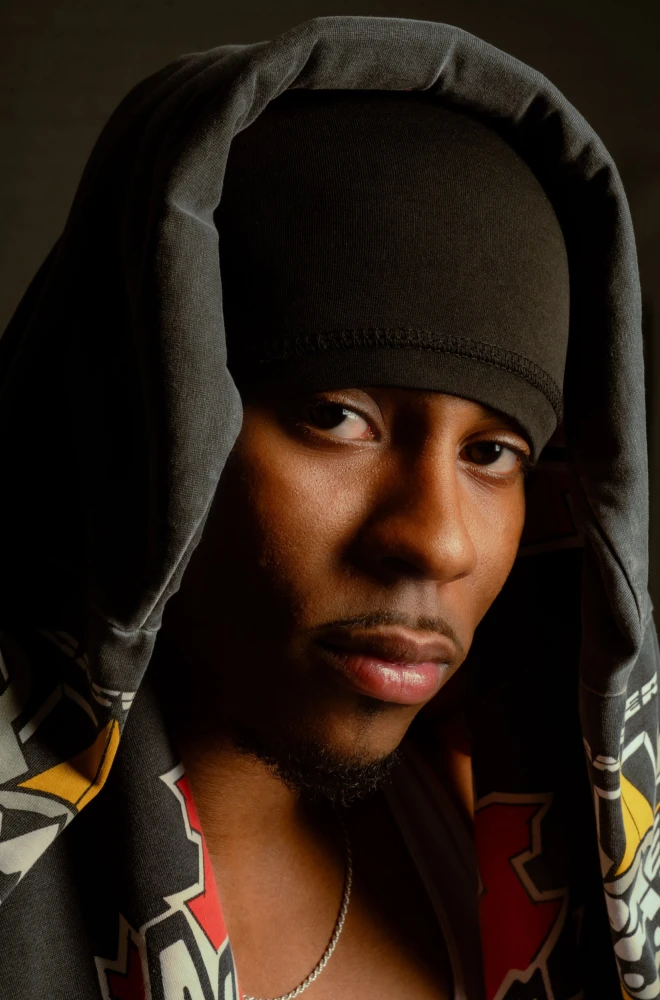 Young artist wearing a gray hoodie, looking to the side with a contemplative expression, silver necklace visible against a dark, shadowy background.