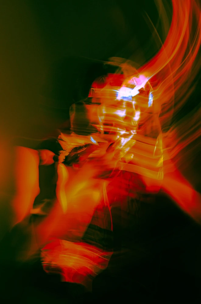 Blurred motion of a figure in intense red and orange hues, creating a dynamic, abstract representation of movement and energy with streaks of light and distorted shapes.