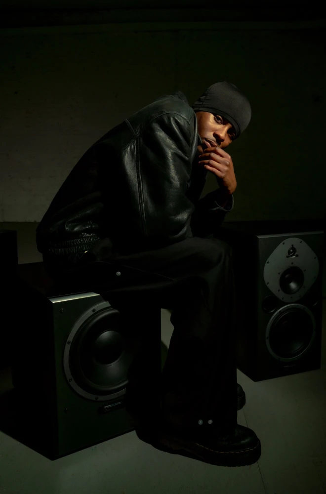 Hooded figure sits pensively between large speakers in a dimly lit, moody setting, with dramatic shadowing highlighting a contemplative pose.