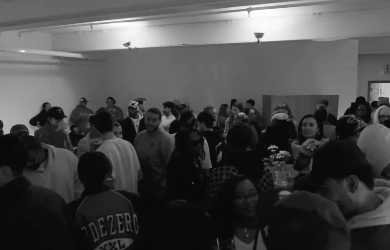 Crowded indoor event space filled with people, some standing in clusters, wearing casual clothing, with dim lighting creating a monochrome atmosphere typical of an underground music venue or art space.