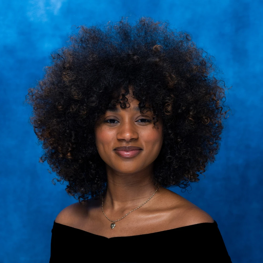 Young woman with voluminous, curly afro hairstyle, wearing a black off-shoulder top, smiling softly against a blue background with a delicate necklace.