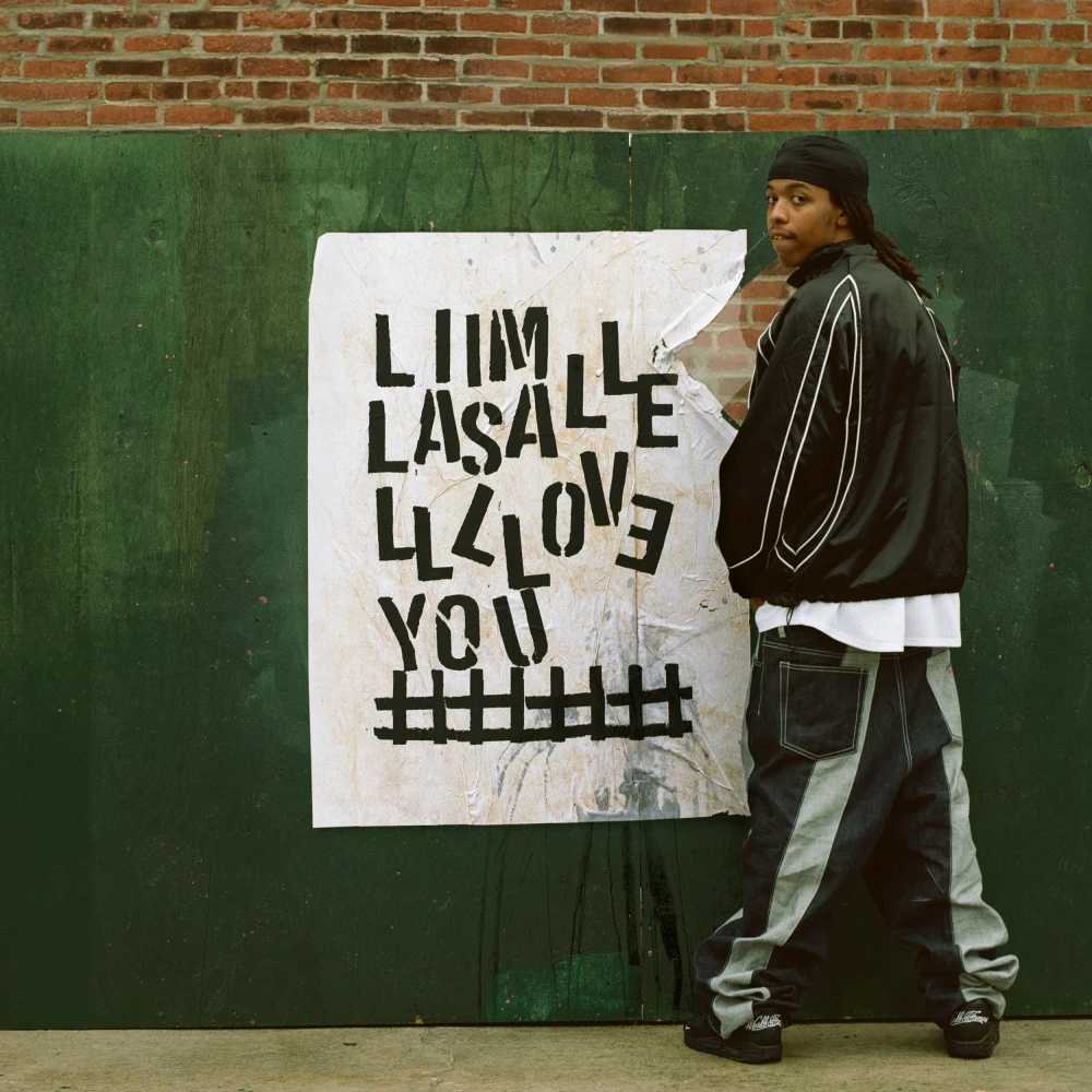 Person in a leather jacket and baggy pants standing next to a handmade poster with distorted text against a green and brick wall background, embodying an urban street style aesthetic.