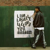 Person in a leather jacket and baggy pants standing next to a handmade poster with distorted text against a green and brick wall background, embodying an urban street style aesthetic.