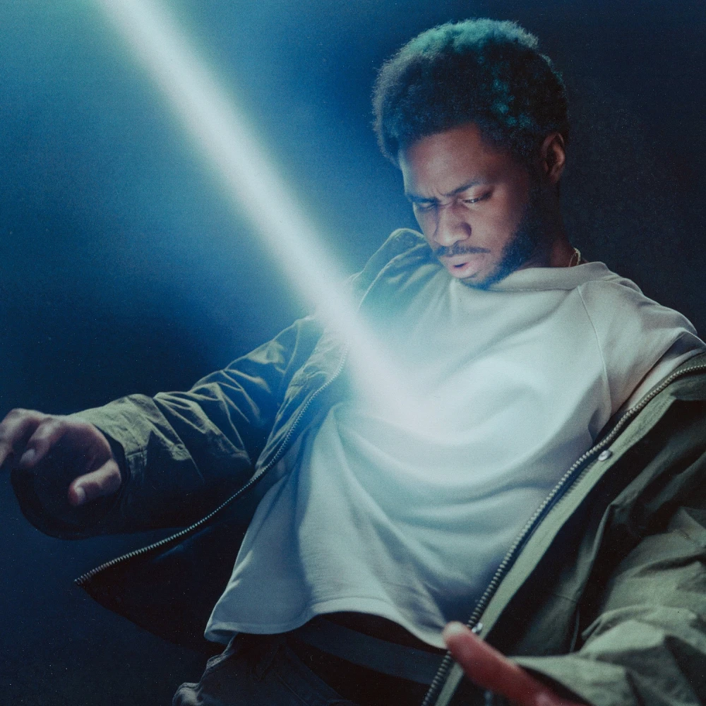 A musician stands illuminated by a bright blue-white light, wearing a white top and jacket, with a contemplative expression in a moody, atmospheric setting.