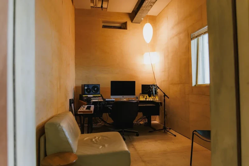 Compact home studio with warm, yellow-toned walls featuring a desk with computer monitors, speakers, and a standing lamp, alongside a minimalist green chair in the foreground.
