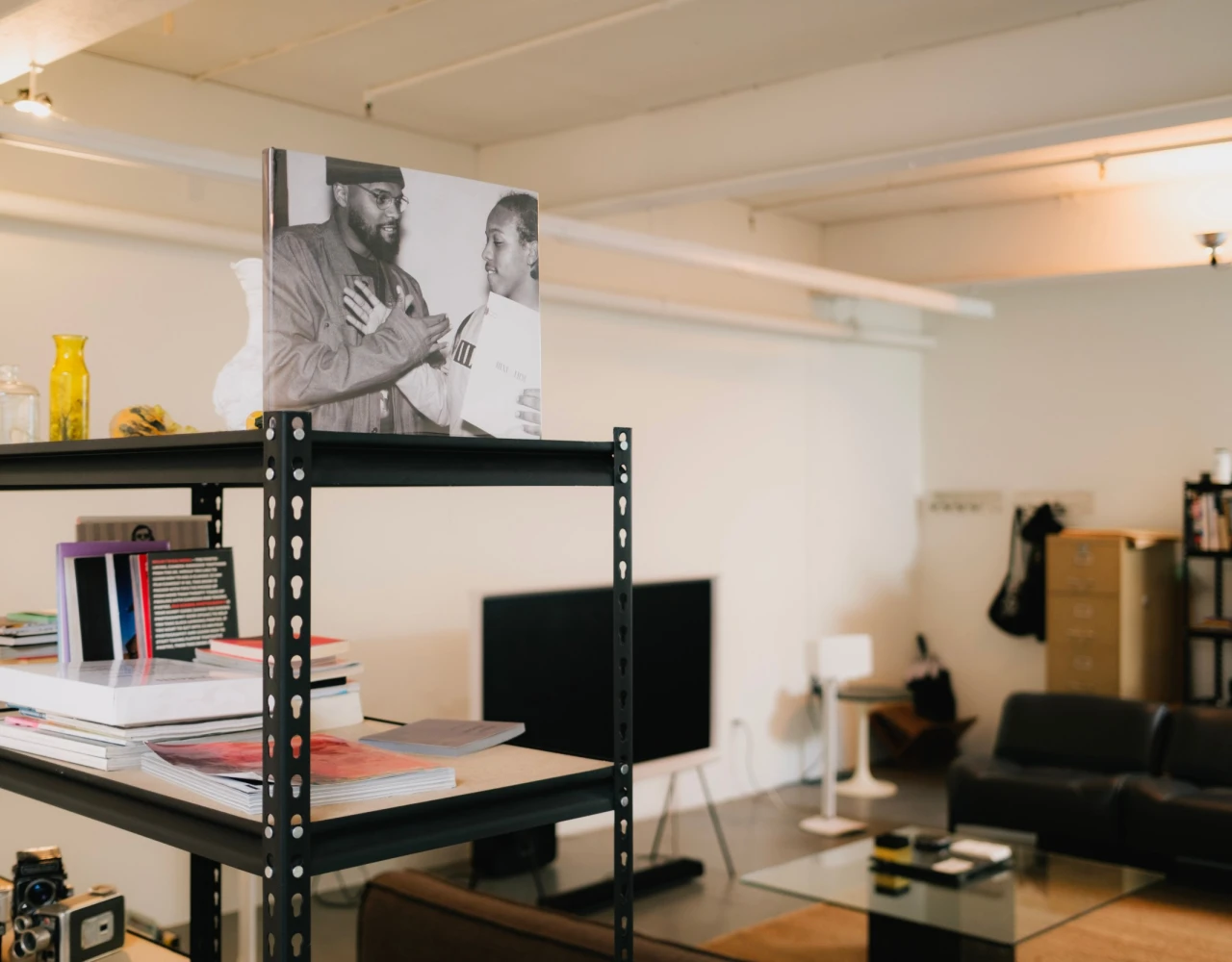 Industrial metal shelving unit in a minimalist workspace with a black and white photographic mural of two people, bookshelves, and a dark sofa in the background, creating an urban creative studio atmosphere.