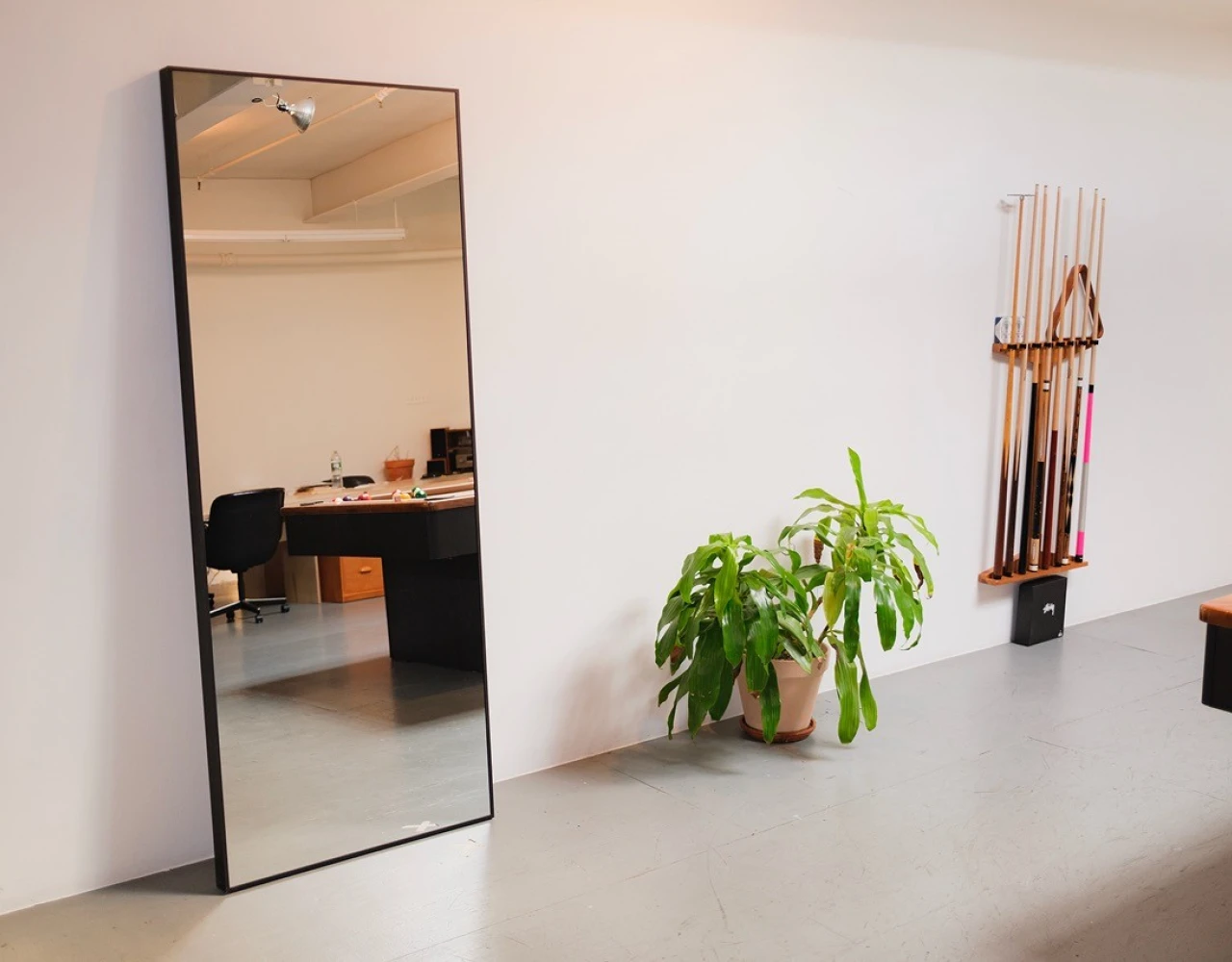 Full-length black-framed mirror leaning against a white wall next to a green potted plant and a wall-mounted rack holding pool cues in a minimalist, light-filled room.