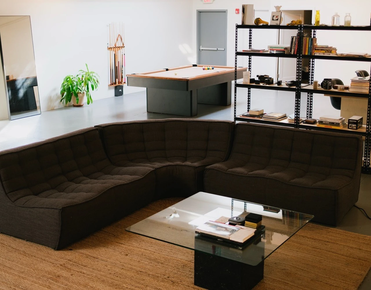 Dark brown sectional sofa in a minimalist studio space with wooden floor, glass coffee table, industrial shelving, and a large mirror leaning against the wall. Green plant adds a touch of color to the neutral, modern interior design.