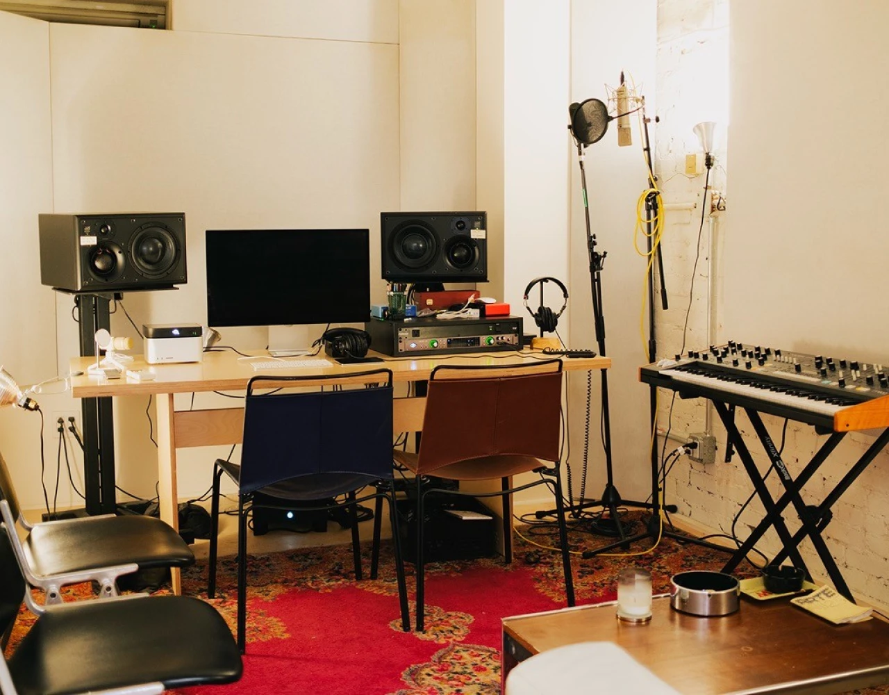 Music production studio with computer workstation, studio monitors, keyboard synthesizer, mixing equipment, microphones, and studio lighting on a red rug in a minimalist white room.
