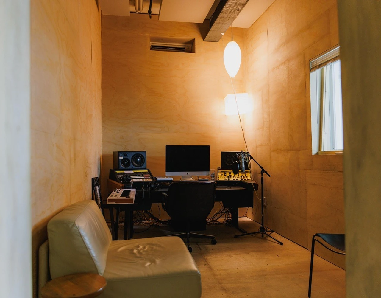 Compact home studio with warm yellow walls, featuring a desk with computer monitors, a microphone, and a floor lamp. Green couch and minimalist decor create an intimate workspace for music production.