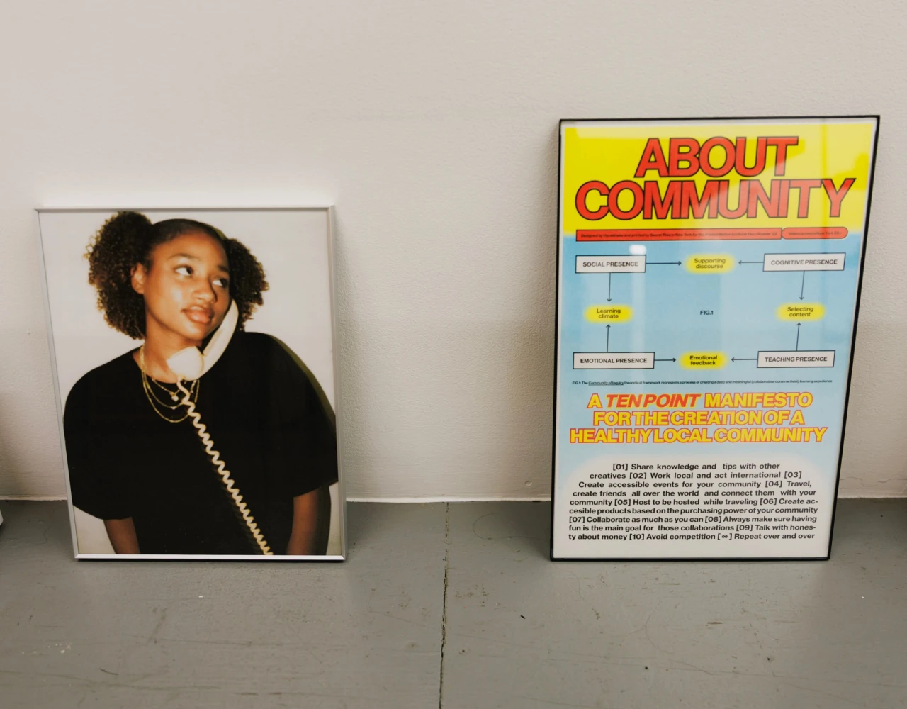 A framed portrait of a smiling person wearing a black top and a necklace, positioned next to a yellow and blue community information poster on a gray surface.