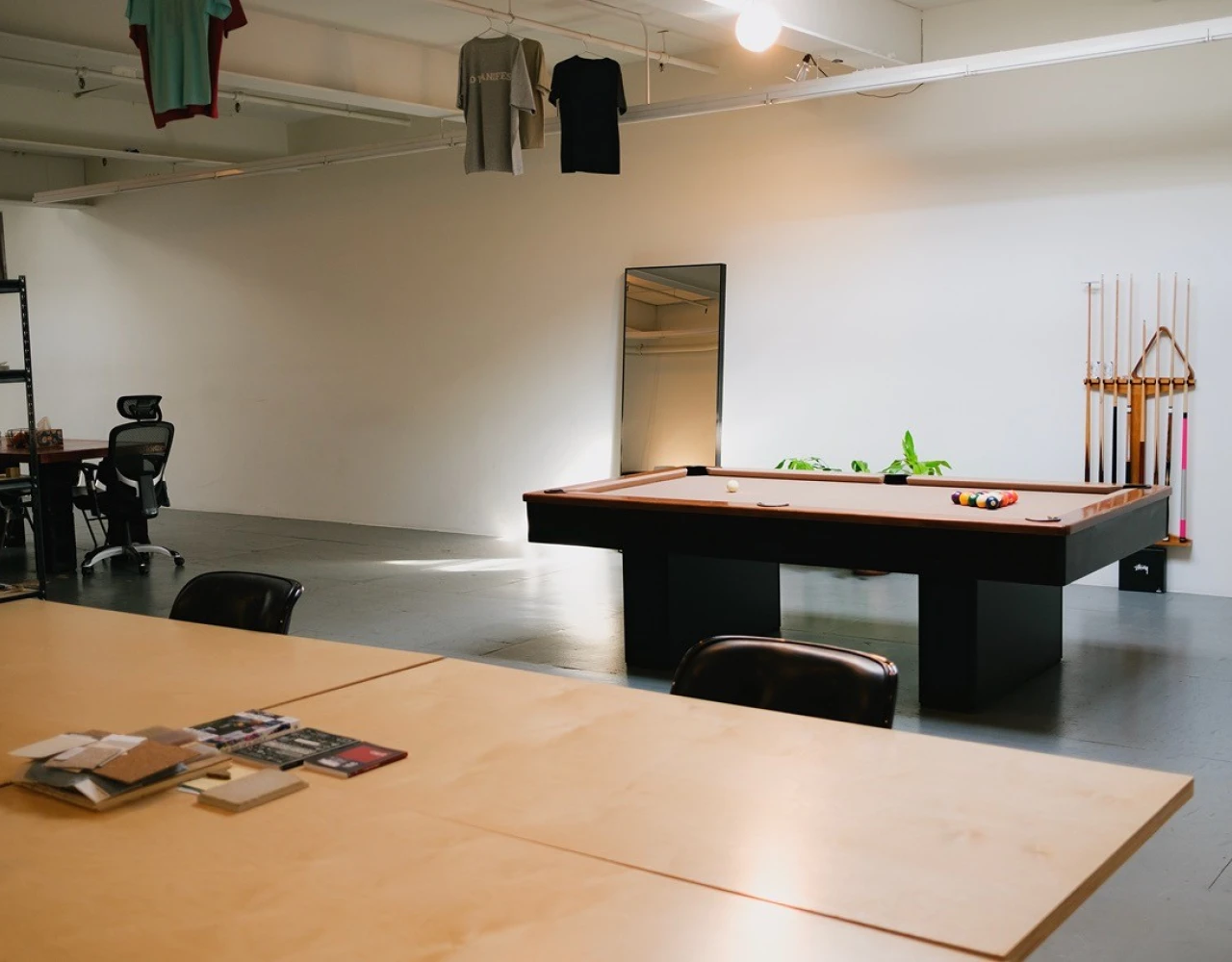 Minimalist studio space with a pool table, wooden conference table, hanging clothes, and office chairs, creating a casual, creative workspace environment.