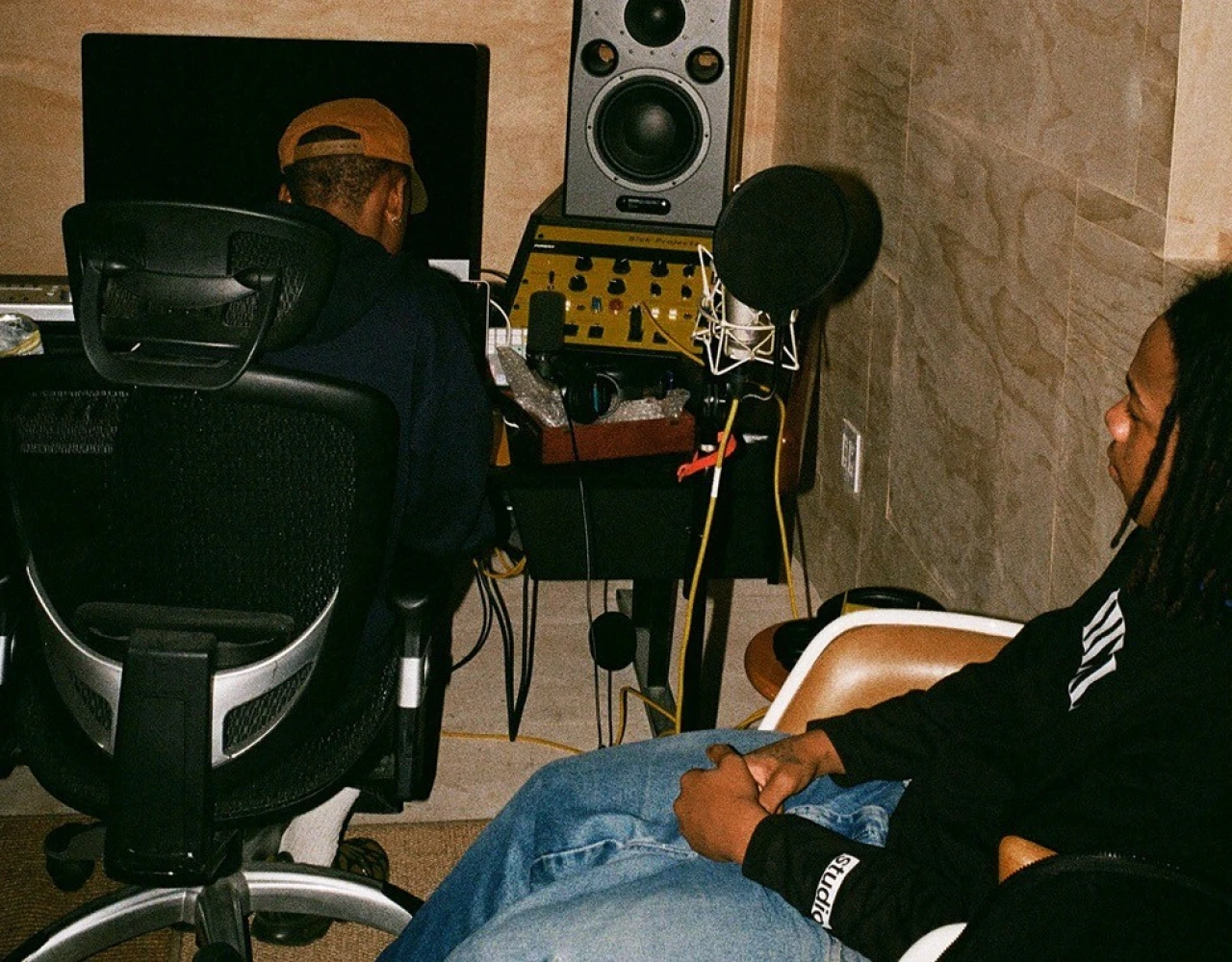 Recording studio scene with music production equipment, including speakers, a microphone, and two people seated in office chairs, focused on audio work.