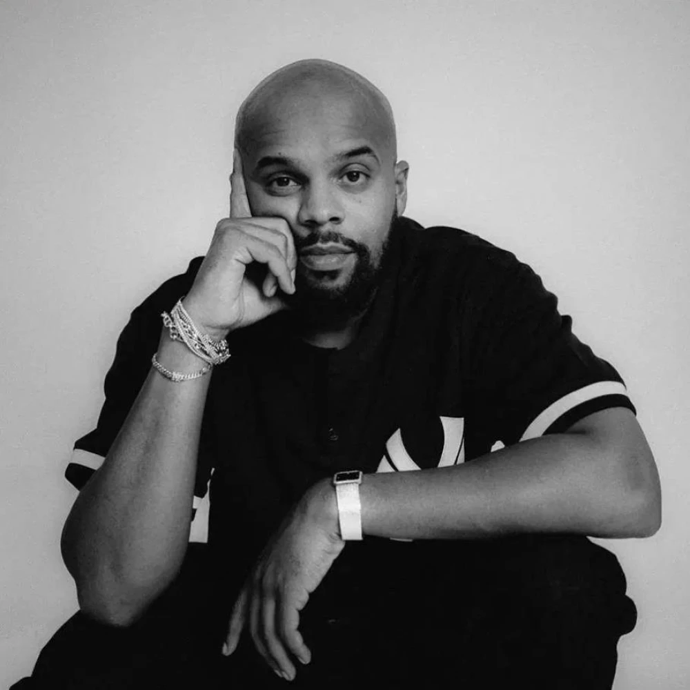Man in a black shirt resting his head on his hand, wearing bracelets and looking contemplatively at the camera in a black and white portrait.