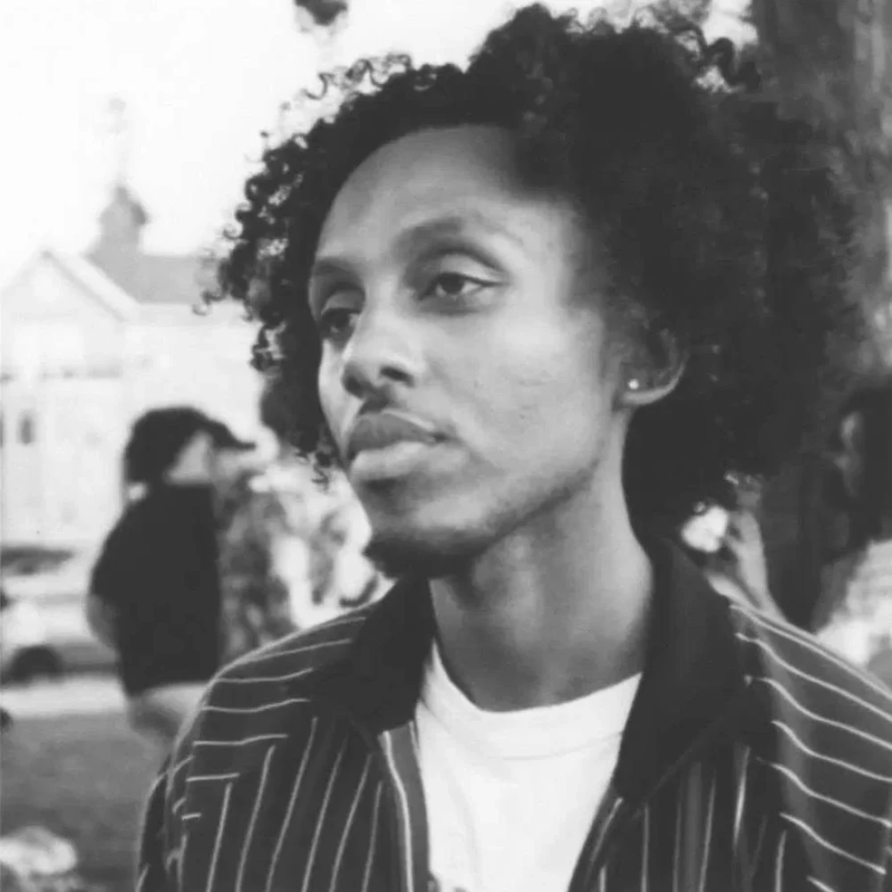Young person with curly hair wearing a pinstriped jacket and white shirt, looking contemplatively to the side with an urban background behind him.