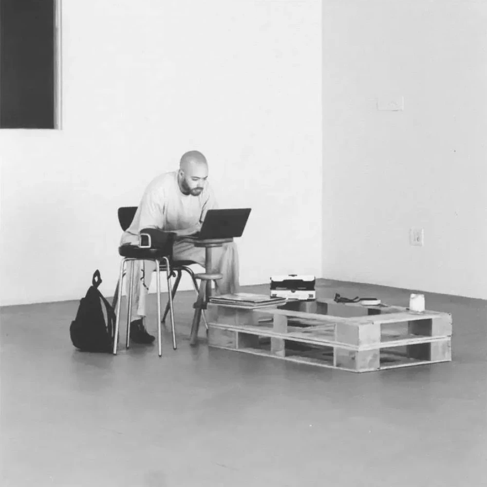 Man works on a laptop at a minimalist workspace with wooden pallets serving as a low table, a black bag nearby, against a white wall and floor.